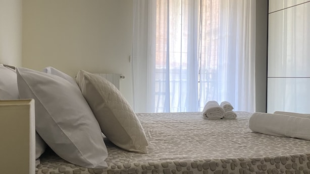 Een slaapkamer met een bed, kussens, een nachtkastje en een raam met dunne gordijnen.