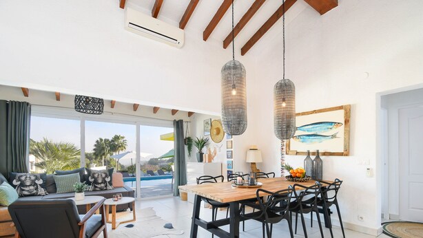 A spacious living area with a wooden dining table, black chairs, and a sofa with patterned cushions. There is a large window with a view of palm trees and a swimming pool. - Villa  with private pool in Moraira, Costa Blanca, Spain for 4 persons (Moraira)