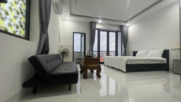 A modern hotel room with a black sofa, a bed with white bedding, a wooden side table, and a large window offering a view of the cityscape. - Moon house villa (Da Nang)