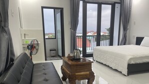 A hotel room with a black leather sofa, a wooden side table with a kettle, a bed with white bedding, and a balcony with a view of buildings. - Moon house villa (Da Nang)