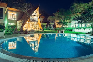 Une piscine dotée d’une architecture moderne, entourée d’une végétation luxuriante et de bâtiments bien éclairés.