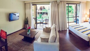 Habitación de hotel con sofá, cama con manta estampada, televisor montado en la pared, zona de comedor con sillas y mesa, y vistas al exterior a través de amplias ventanas.