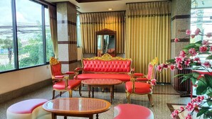 A hotel lobby with a red and gold sofa set, a round table, and matching chairs. There is a large mirror above the sofa and a potted plant with pink flowers to the right. - The Popular place (Bang Phli)