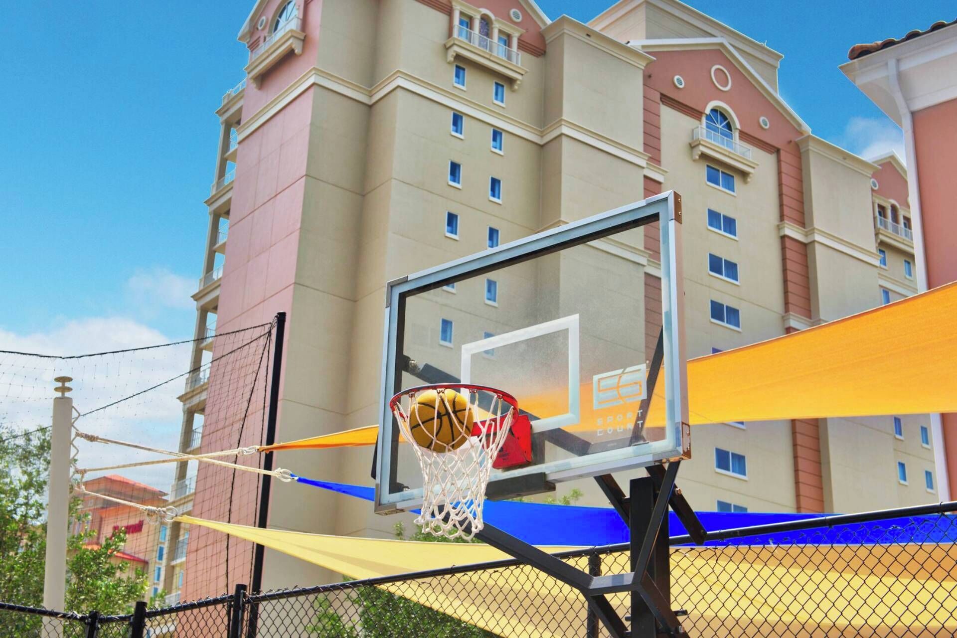 A basketball hoop with a net, a chain-link fence, and a building with multiple windows in the background.