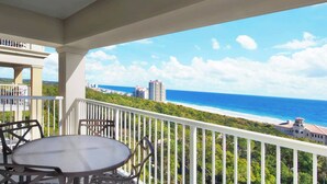 A balcony with a table and chairs, overlooking a beach and ocean with buildings in the distance.
