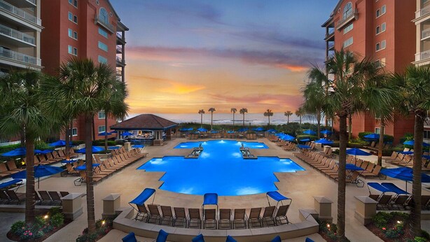 A resort pool area with multiple pools, lounge chairs, and umbrellas, surrounded by palm trees and a sunset view.
