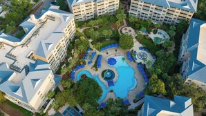 An aerial view of a hotel complex with multiple buildings, a central swimming pool, and surrounding greenery.
