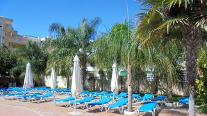 Zona de piscina amb gandules blaves i ombrel·les blanques, envoltada de palmeres i un paviment de rajola.