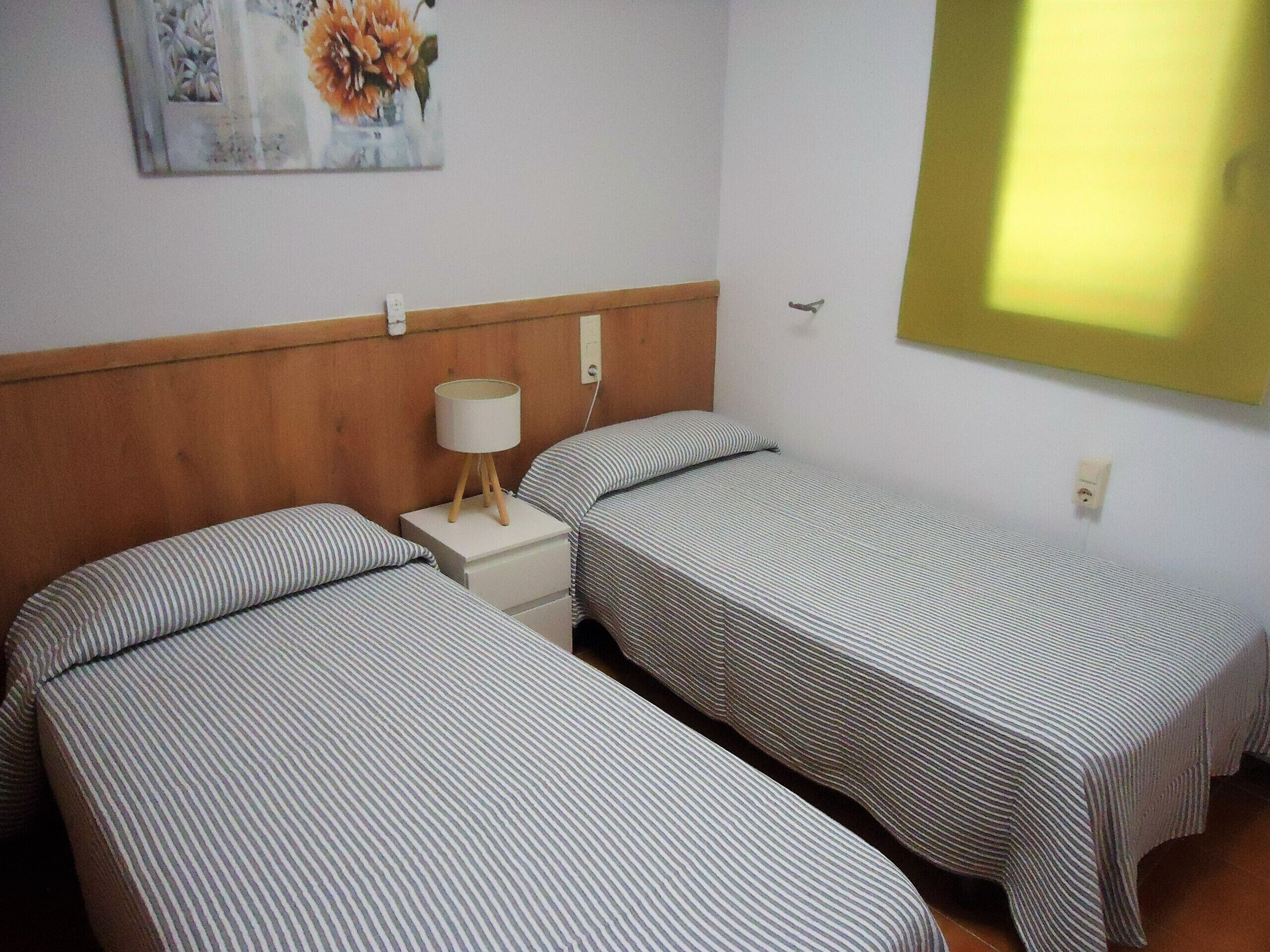 A room with two single beds, a nightstand with a lamp, a wall-mounted power outlet, and a painting of flowers.