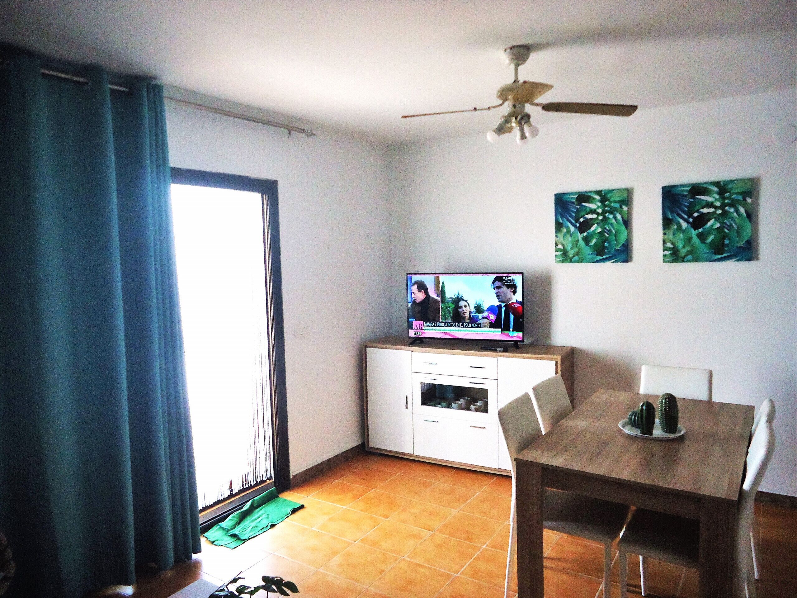 A room with a TV on a cabinet, a ceiling fan, a dining table with chairs, and a window with curtains.