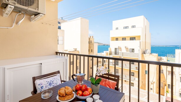 Un balcón con mesa lista para el desayuno, una planta en maceta y vistas al mar y edificios.