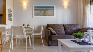 Een woonkamer met een bruine bank, witte stoelen, een kleine tafel en een schilderij van een strandgezicht.