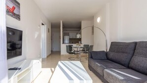 A modern living room with a grey sofa, a white coffee table, and a flat-screen TV on a white stand. There is a dining area with a table and chairs, and a kitchen in the background. - Michelangelo by Interhome (Salou)