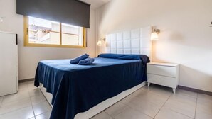 A bedroom with a bed, a nightstand, a window with blinds, and a wall-mounted lamp. - Miramar Nautic by Interhome (Salou)