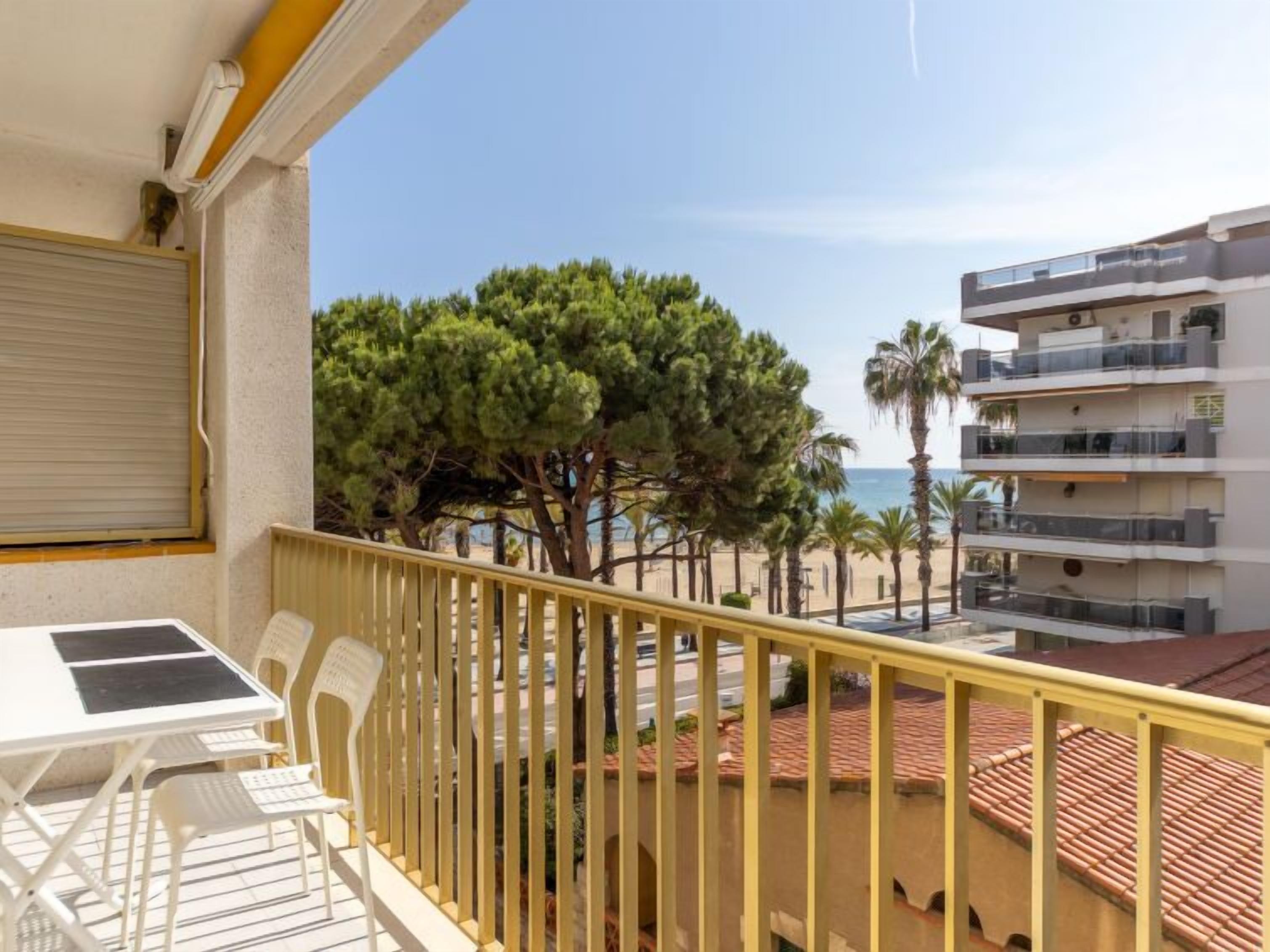 A balcony with a table and chairs, overlooking a beach with palm trees and a building with balconies.