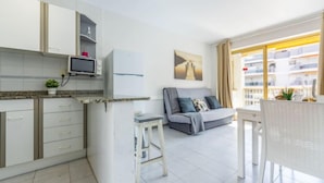A modern kitchen with white cabinets, a built-in microwave, and a small island with stools. There is a grey sofa with cushions in the living area, and a large window with curtains. - Miramar Nautic by Interhome (Salou)