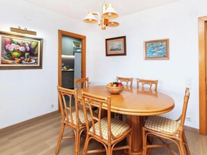 Un comedor con una mesa redonda de madera, seis sillas de mimbre con cojines a cuadros, y un frutero. Hay tres obras de arte enmarcadas en las paredes y se ve una cocina a través de una puerta abierta.