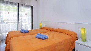 Ein Hotelzimmer mit zwei Betten, weißen Wänden, einem Fenster mit leichtem Vorhang und einem Nachttisch mit Lampe.