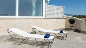 Terraza en la azotea con dos tumbonas, una parrilla y un amplio ventanal con vistas a un paisaje pintoresco.