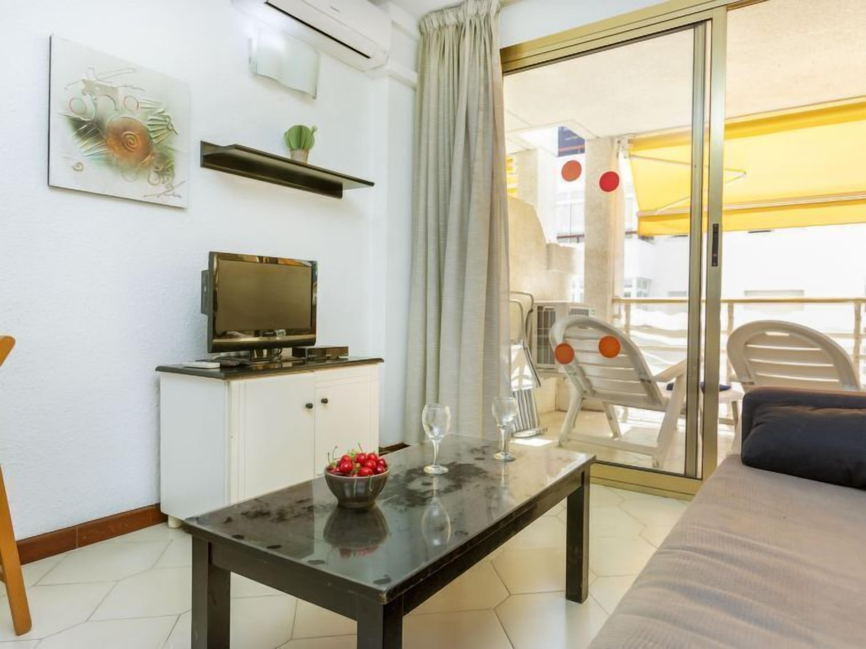 Une chambre moderne avec un téléviseur posé sur un meuble, une table à manger agrémentée d’un saladier de fruits et de verres à vin, et un balcon avec des chaises et un parasol jaune.