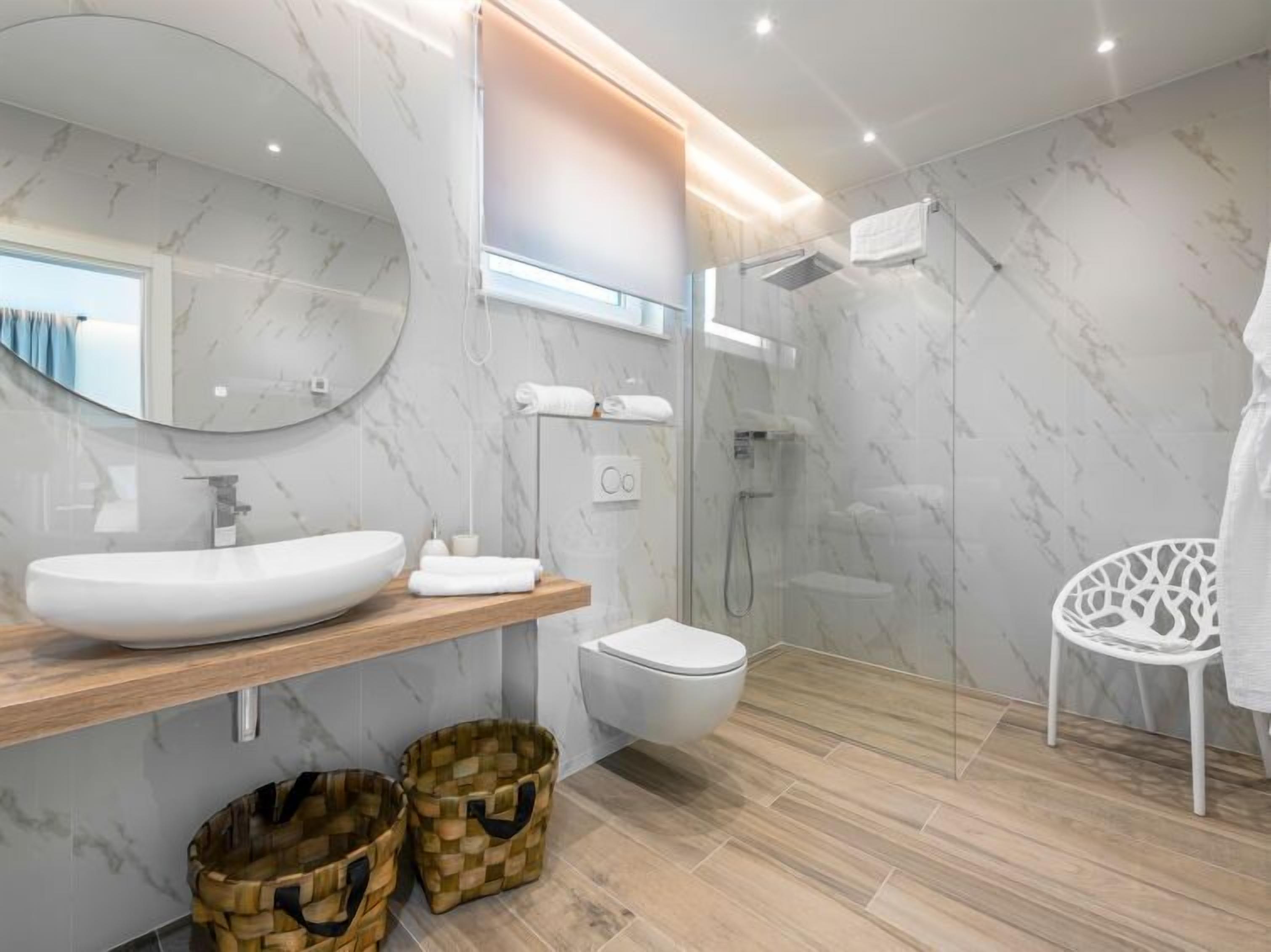 A modern bathroom with a large oval sink, a toilet, a glass-enclosed shower, and a white chair.