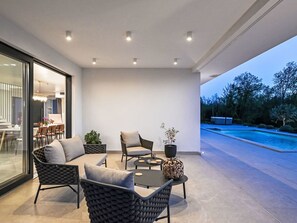 A modern outdoor living area with a pool, furnished seating, and a dining area visible through a glass wall.