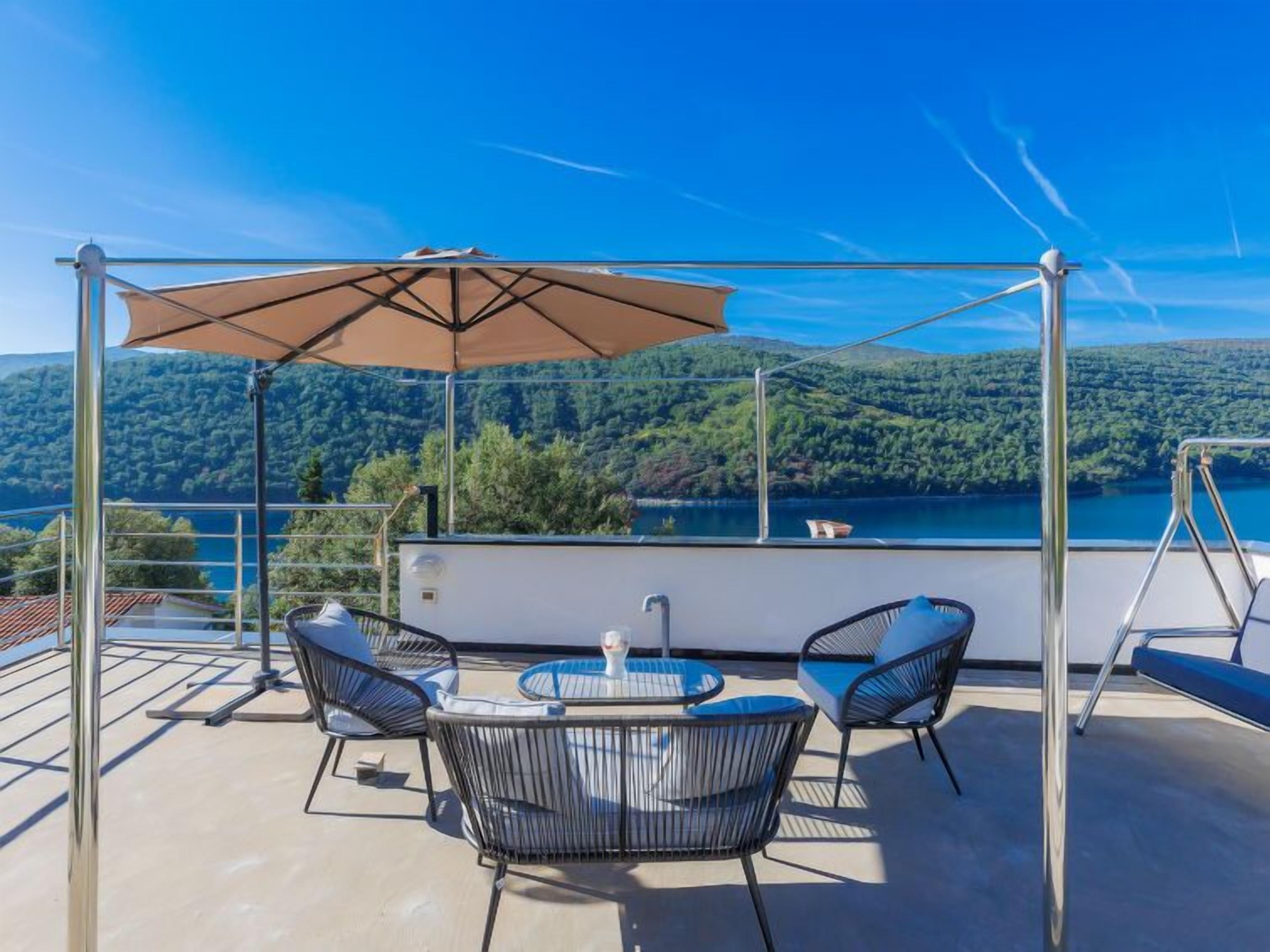 A terrace with a table and chairs, a large umbrella, and a scenic view of a lake and mountains.