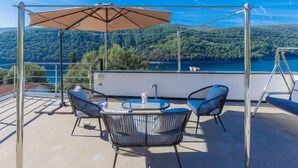 Una terraza con una mesa y sillas, un paraguas grande y una vista panorámica de un lago y montañas.