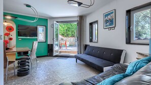 Una sala moderna con un sofá de cuero negro, un área de comedor con pared verde y un televisor, y vista a zonas verdes afuera.