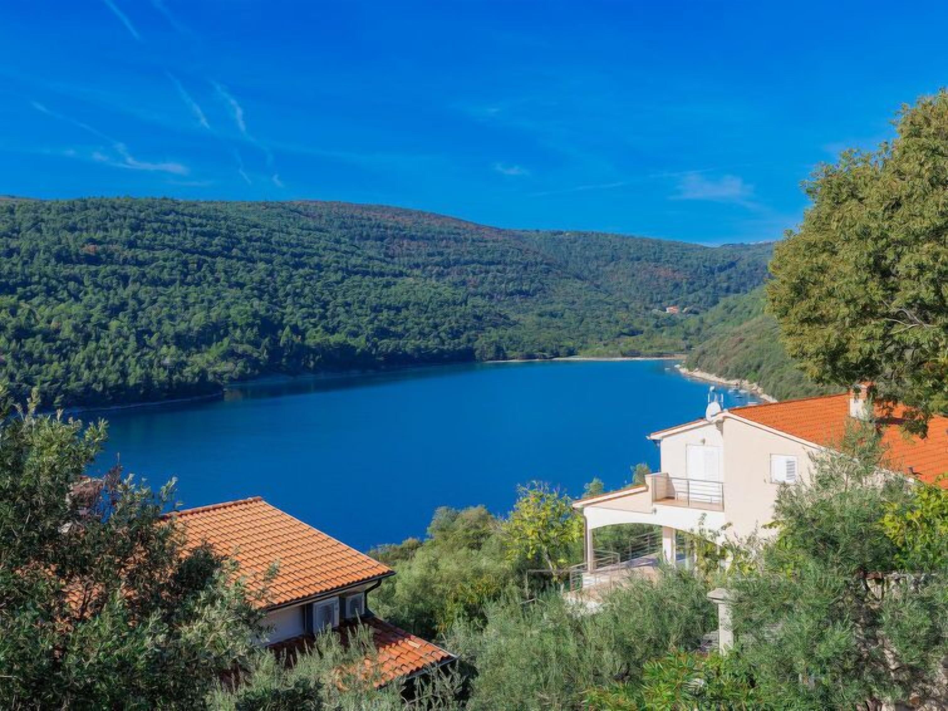 A coastal house with a red-tiled roof, surrounded by lush greenery and overlooking a calm blue lake.
