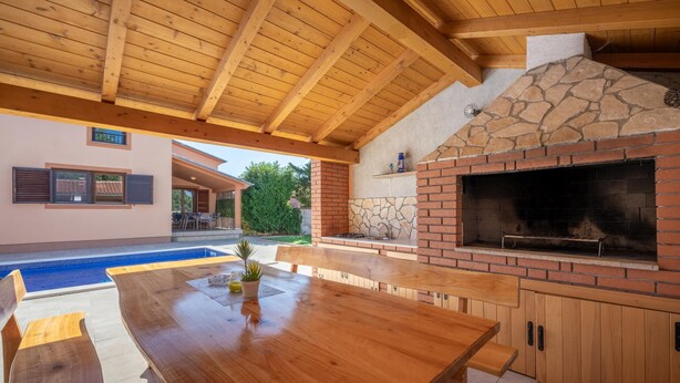 Una veranda esterna con tavolo e panche in legno, un caminetto in pietra e una piscina.