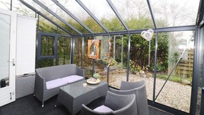 Ein Wintergarten mit Korbmöbeln, einem Glastisch und Blick auf einen Garten.