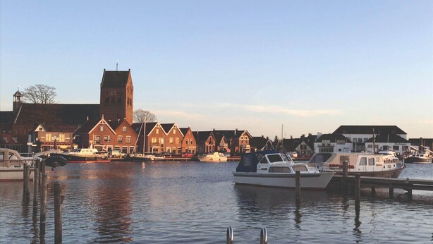 Ein Hafen mit vertäuten Booten, eine Kirche mit einem hohen Turm und traditionelle Gebäude entlang der Uferlinie.