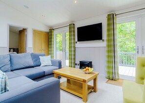 A living room with a sofa, a coffee table, a flat-screen TV, and a balcony with green curtains. - Newton Brook Elegance Lodge (Newton Saint Loe, Nr Bath)