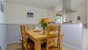 A dining area with a wooden table, chairs, a vase with flowers, a kitchen in the background, and a painting on the wall.