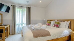 A well-lit bedroom with a large bed, a bedside table, a desk, and a TV mounted on the wall.