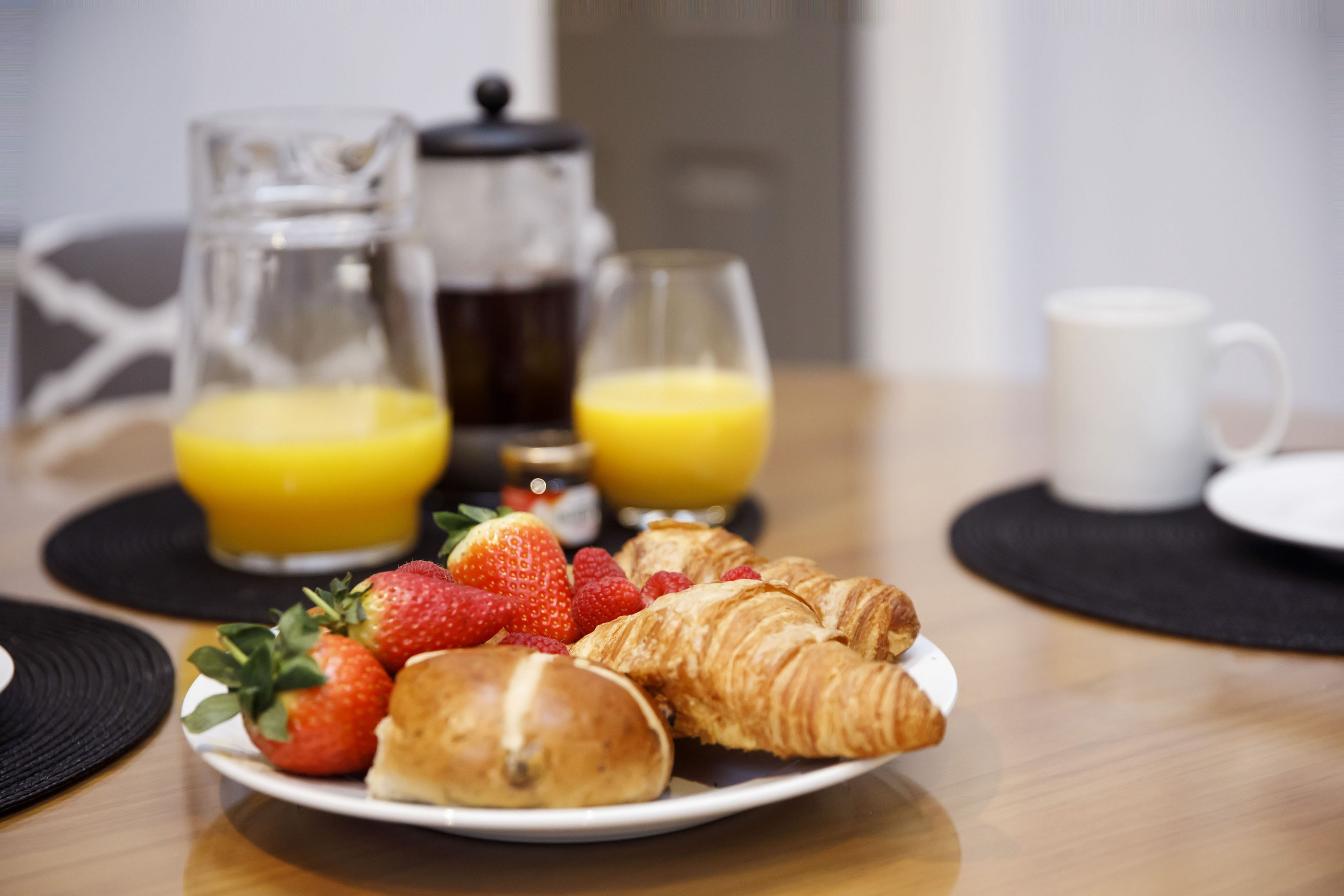 A breakfast setting with croissants, strawberries, and a glass of orange juice on a plate, with a coffee pot and mug in the background.