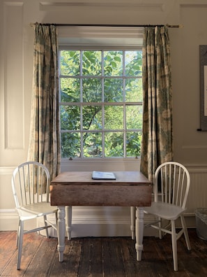 A room with a window, two white chairs, a wooden table, and a framed picture on the wall.