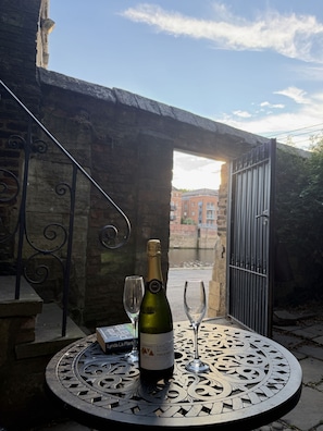 A table with a bottle of wine and two glasses, set against a brick wall with a metal gate and a view of buildings in the background.