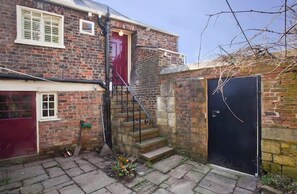 Traditional brick building with a red door and black door, featuring stone steps and a small window with shutters.