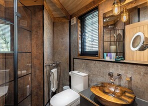 Ein modernes Badezimmer mit einem Holzboden, einer Toilette, einer Duschkabine aus Glas und einem Fenster mit Jalousien.