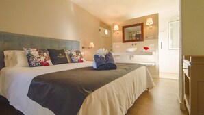 Habitación de hotel con una cama grande, una mesita de noche, un baño con lavabo y espejo, y un escritorio de madera.