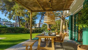 Un patio exterior con una mesa de madera dispuesta para comer, rodeado de sillas de mimbre y una cesta colgante.