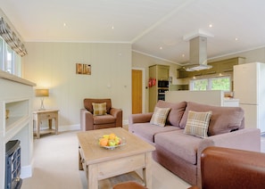 A cozy living room with a sofa, a small table with fruit, a chair, and a kitchen area in the background.