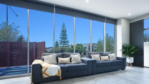 A modern living room with a grey sofa, white and yellow throw pillows, and a large window offering a view of trees and a fence.