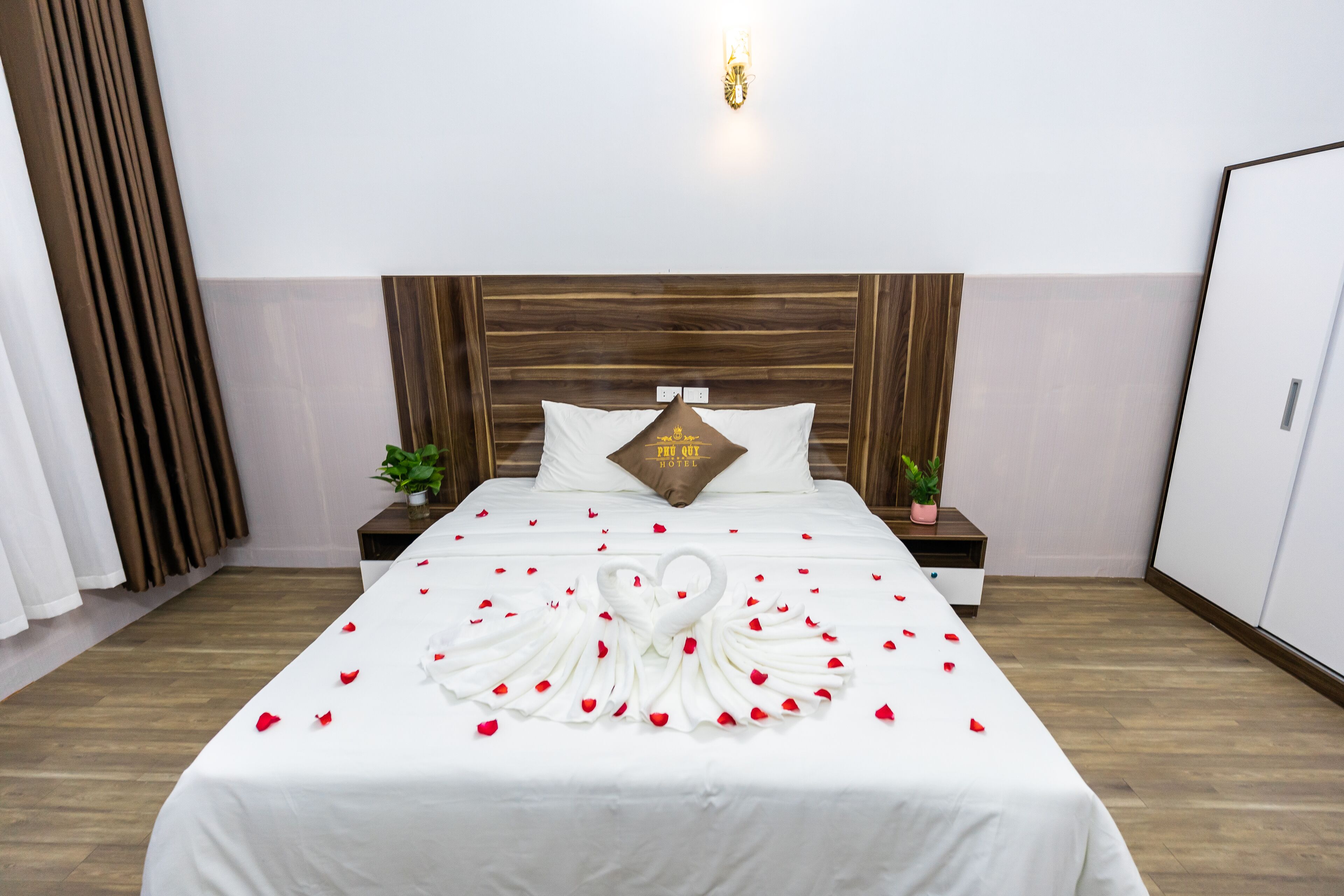 Een hotelkamer met een net opgemaakt bed, een houten hoofdeinde, een nachtkastje met een plant en een wandlamp.