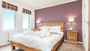 A bedroom with a wooden bed, two bedside tables, a window with a plaid blind, and a wall-mounted lamp. - Netherfield Watersedge Lodge (Newton Saint Loe, Nr Bath)