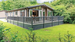 A single-story house with a deck, surrounded by lush greenery. - Netherfield Watersedge Lodge (Newton Saint Loe, Nr Bath)
