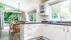 A modern kitchen with white cabinets, a dining area with wooden chairs, and large windows offering a view of greenery. - Netherfield Watersedge Lodge (Newton Saint Loe, Nr Bath)