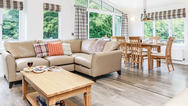 A modern living room with a sofa, dining table, and chairs, large windows with curtains, and a wooden floor. - Netherfield Watersedge Lodge (Newton Saint Loe, Nr Bath)